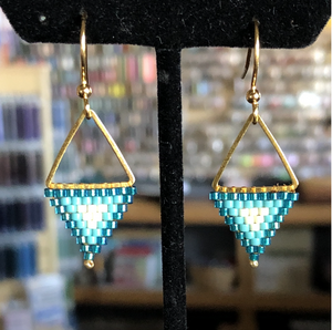 Gold and turquoise  seed bead beaded triangle earrings on a black stand with a blurred background.