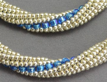Close-up of a beaded strand or bracelet with rows of small metallic silver beads and a central band of glossy blue faceted beads, shown on a gray textured background.