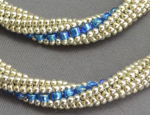 Close-up of a beaded strand or bracelet with rows of small metallic silver beads and a central band of glossy blue faceted beads, shown on a gray textured background.