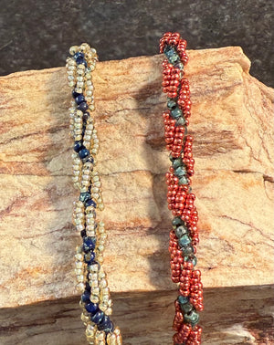 Two handmade beaded bracelets displayed on a textured stone slab. The bracelet on the left features cream-colored seed beads with dark blue accents, and the bracelet on the right features red seed beads with green and blue accents.